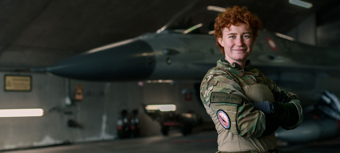 En ung kvindelig flymekaniker med kort rødt hår poserer i uniform. I baggrunden ses et F16-fly i en hangar.