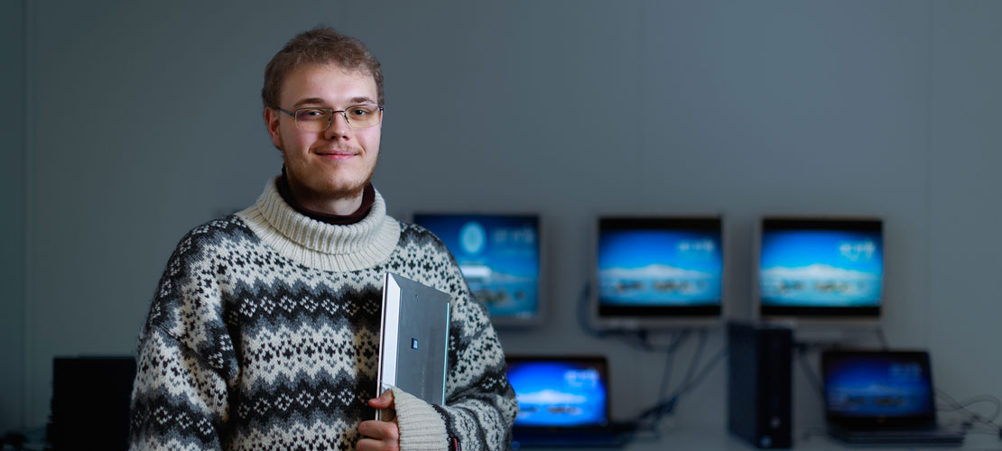En ung mand med lyst hår, uldsweater og briller poserer. Han har en bærbar computer i den ene arm. I baggrunden ses en række tændte computerskærme