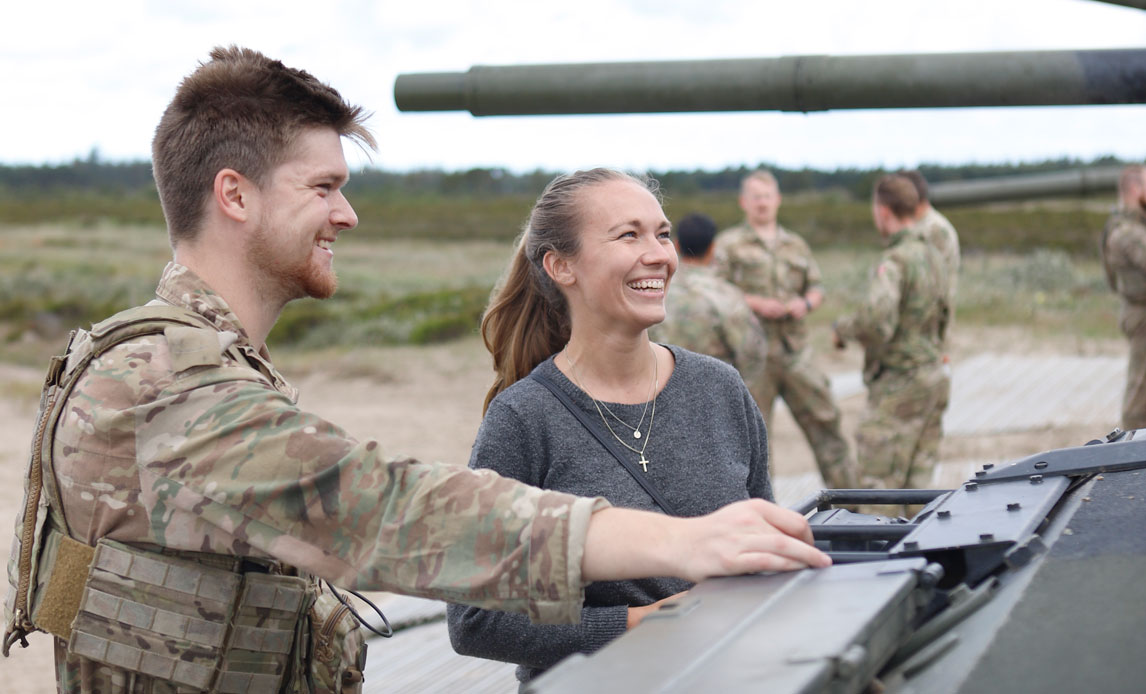 En mandlig soldat i uniform og en kvinde i civilt tøj står ved en kampvogn. De smiler begge og kigger på noget uden for billedet