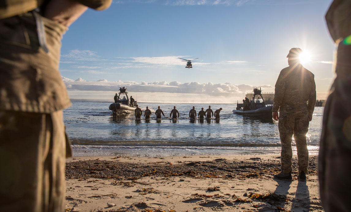 En række af frømænd på vej op ad vandet på en strand. På stranden kan i forgrunden ses 3 soldater med ryggen til. Bag soldaterne i vandet kan ses to både og en helikopter. Der er blå himmel og solskin.