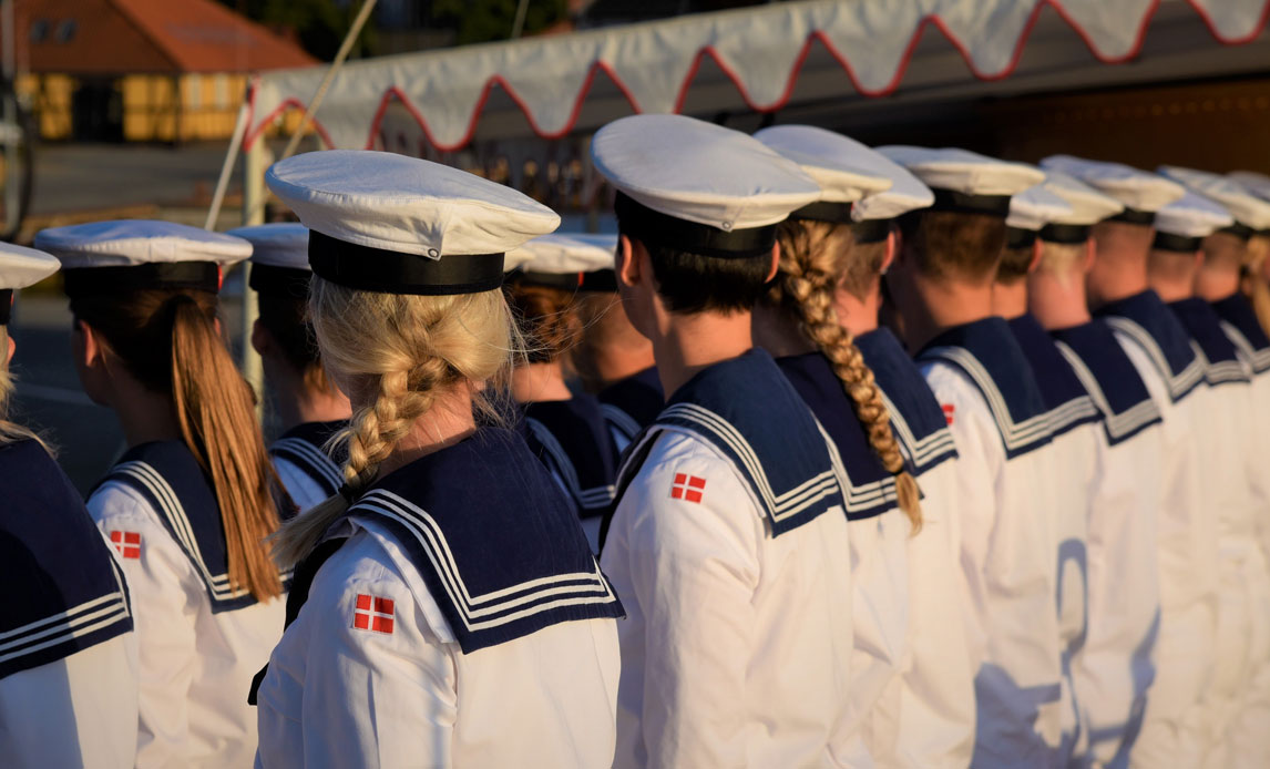 Værnepligtige (både mænd og kvinder) fra Søværnet står i geledder med ryggen til kameraet. De har hvide uniformer med blå krave på og kasketter.