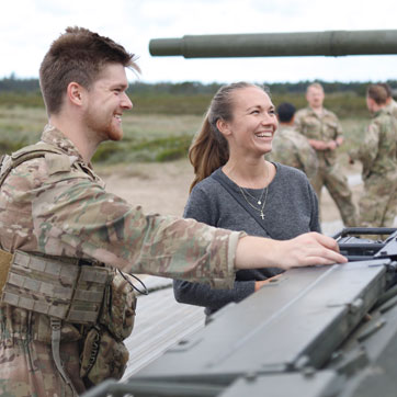En mandlig soldat i uniform og en kvinde i civilt tøj står ved en kampvogn. De smiler begge og kigger på noget uden for billedet